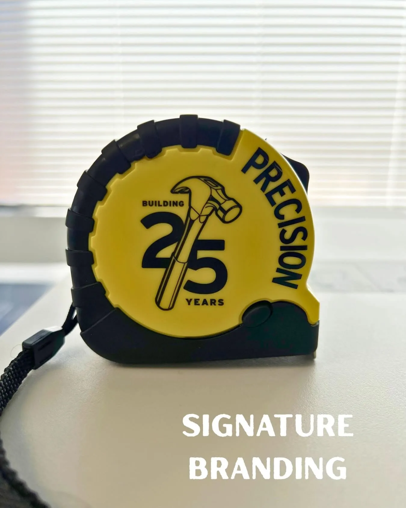 Close-up of a custom-branded yellow-and-black tape measure printed with a 'Building 25 Years / Precision' hammer logo, sitting on a desk
