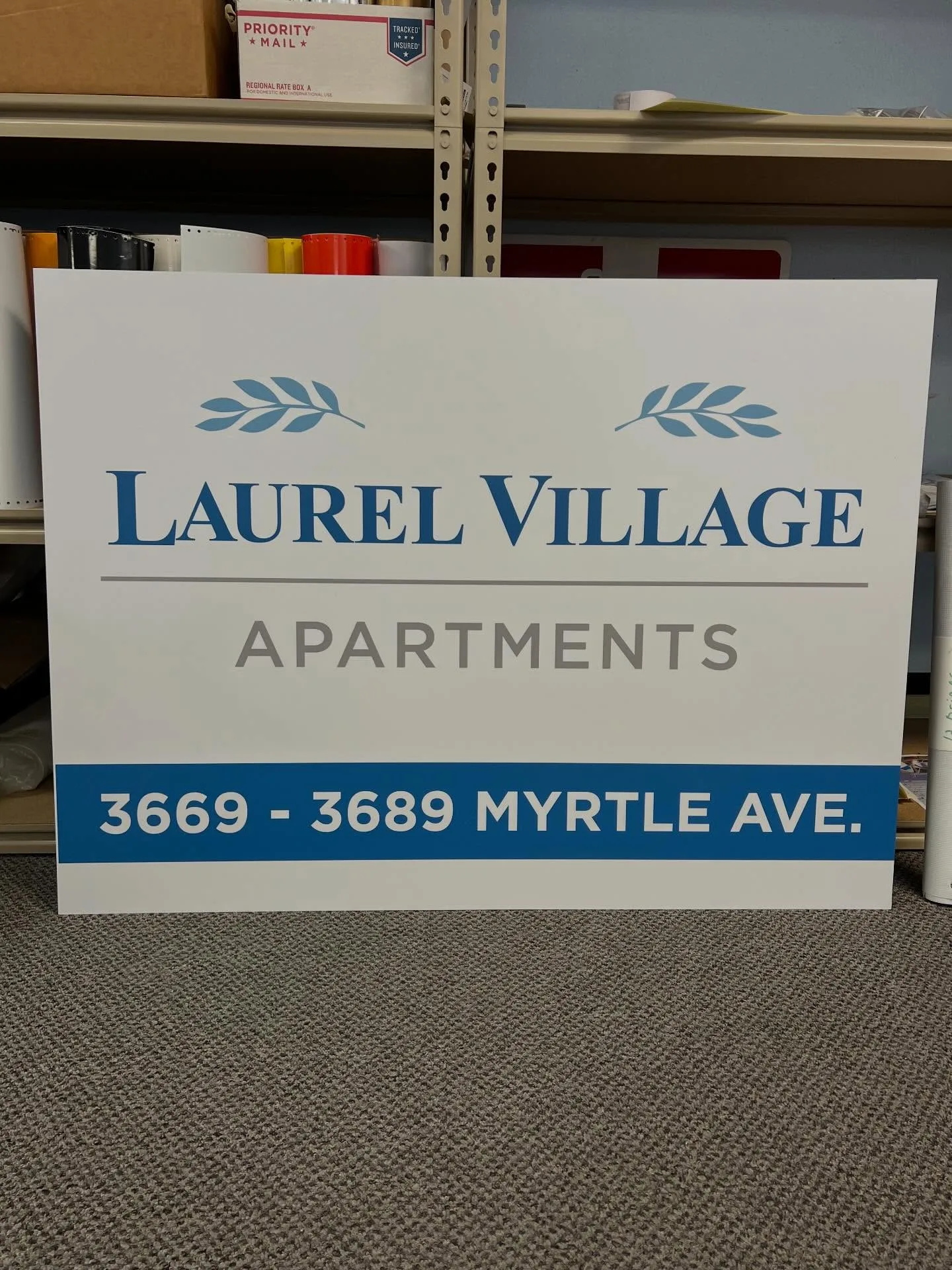 Freshly printed white rigid panel sign for Laurel Village Apartments leaning against shop shelving stocked with vinyl rolls and supplies
